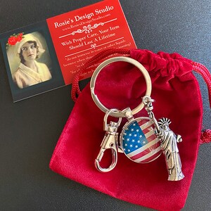 Op de afbeelding: Een zilveren sleutelhanger met een rood, wit en blauwe Amerikaanse vlag charme en een zilveren Vrijheidsbeeld charme. De sleutelhanger zit in een rode fluwelen zak. De tekst op de kaart luidt "Rosie's Design Studio" en "Met de juiste zorg, zal uw item een ​​leven lang meegaan".