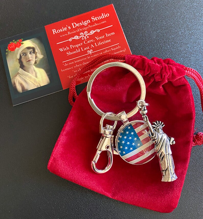 Op de afbeelding: Een zilveren sleutelhanger met een rood, wit en blauwe Amerikaanse vlag charme en een zilveren Vrijheidsbeeld charme. De sleutelhanger zit in een rode fluwelen zak. De tekst op de kaart luidt "Rosie's Design Studio" en "Met de juiste zorg, zal uw item een ​​leven lang meegaan".