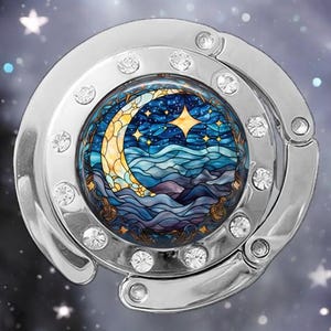 May include: A silver-toned, foldable purse hook with a round, clear crystal-encrusted frame. The frame features a stained glass design of a crescent moon, stars, and waves in shades of blue, yellow, and purple.