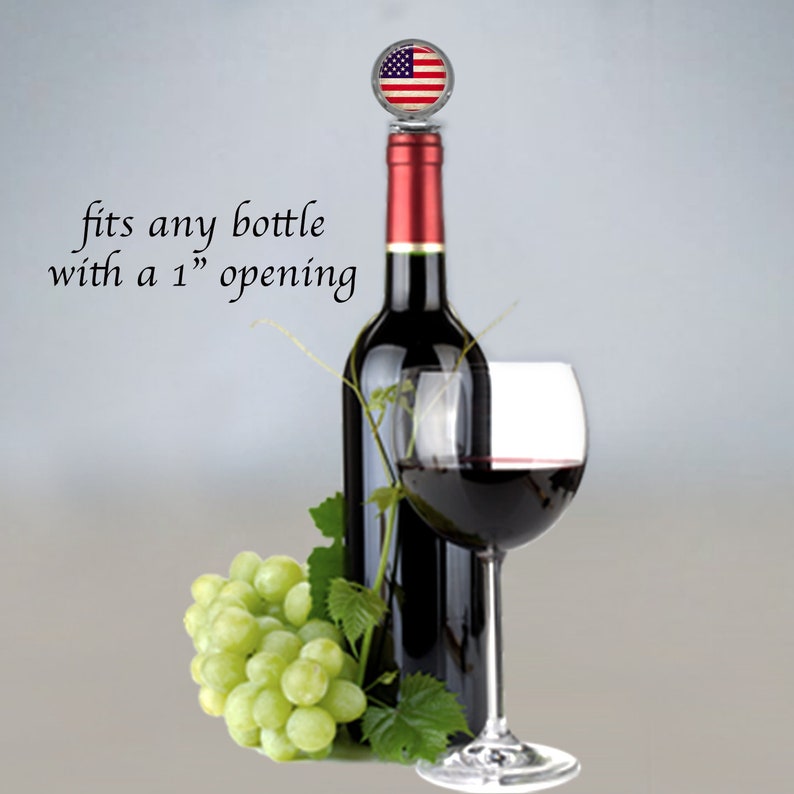 May include: A red wine bottle with a silver bottle stopper featuring an American flag design. The bottle is next to a glass of red wine and a bunch of green grapes. The text "fits any bottle with a 1" opening" is visible in the image.