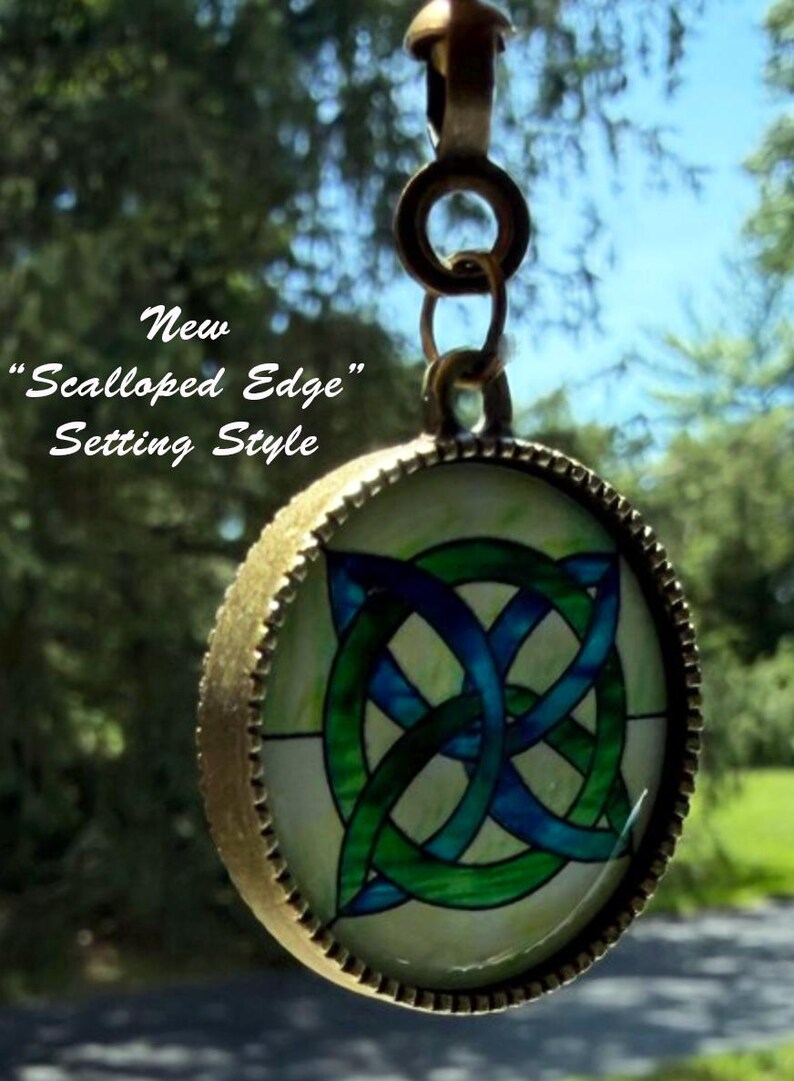 Op de afbeelding: Een ronde hanger met een blauw en groen Keltisch knoopontwerp in een bronskleurig metalen frame met een geschulpte rand. De hanger hangt aan een ronde ring. De tekst op de afbeelding luidt: New "Scalloped Edge" Setting Style.