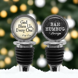 May include: Two silver-toned bottle stoppers with black rubber seals. The left stopper features a cream-colored circular insert with the text God Bless Us, Every One! ~ Tiny Tim. The right stopper has a black patterned insert with BAH HUMBUG ~ Scrooge.