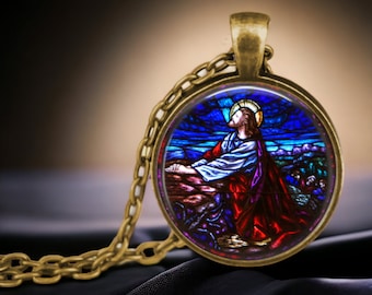 May include: A gold chain necklace with a round pendant featuring a stained glass image of Jesus praying. The stained glass image is set against a blue and purple background with a red robe and a halo.