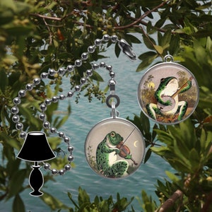 May include: Two round, silver-toned fan pull charms featuring a green frog playing a musical instrument. One frog plays a banjo, the other plays a violin. The charms are hanging from a silver chain.