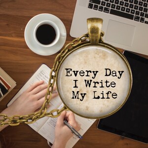 May include: A gold chain necklace with a round pendant featuring a vintage-style background and the text "Every Day I Write My Life".