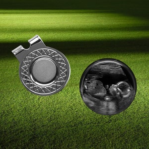 May include: A silver golf ball marker with a black and white patterned design and a black magnetic disc. Next to it is a black and white ultrasound image of a baby.