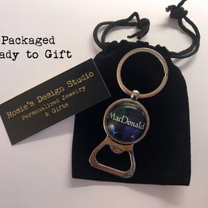 May include: A silver bottle opener keychain with a round, glass charm featuring a green, blue and red tartan pattern and the text "MacDonald". The keychain is packaged in a black velvet bag.