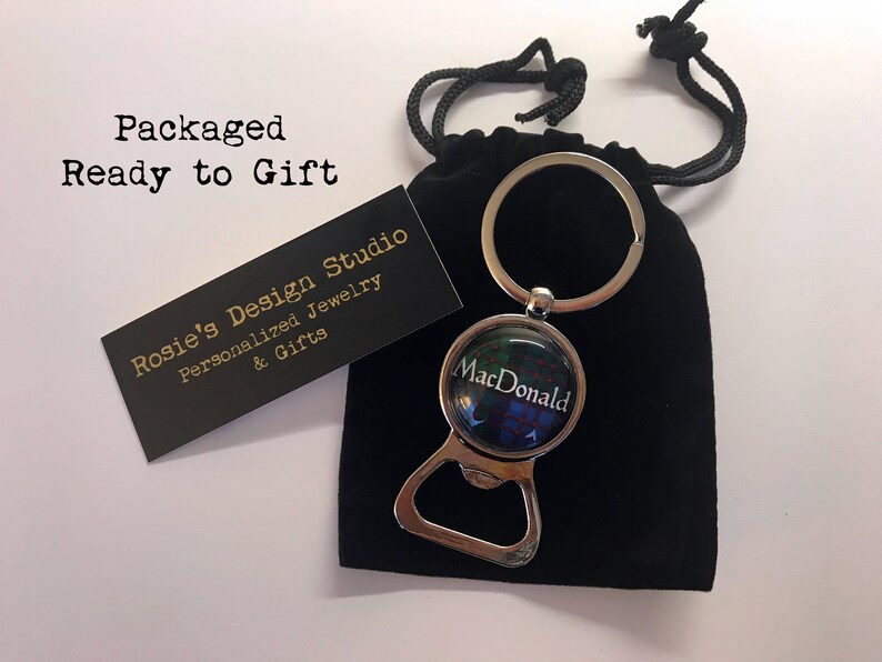 May include: A silver bottle opener keychain with a round, glass charm featuring a green, blue and red tartan pattern and the text "MacDonald". The keychain is packaged in a black velvet bag.