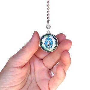 Op de afbeelding: Een ronde trekketting voor een plafondventilator met een zilverkleurig metalen frame en een glazen cabochon-inzetstuk. Het inzetstuk toont een symmetrisch bloemmotief in tinten helderblauw, donkerblauwgroen en wit op een lichtgroene achtergrond.