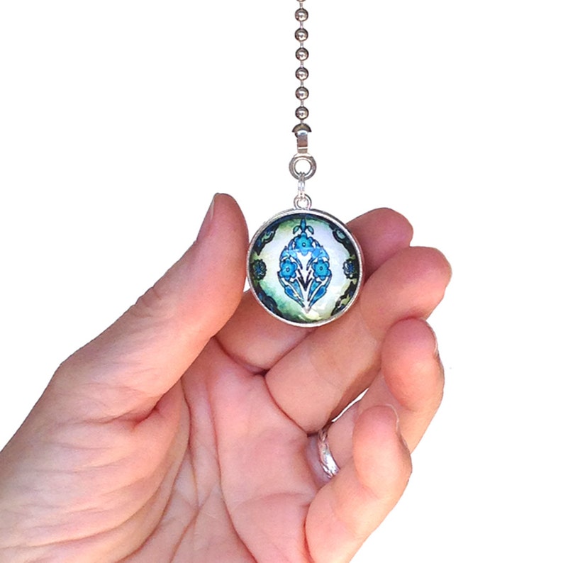 Op de afbeelding: Een ronde trekketting voor een plafondventilator met een zilverkleurig metalen frame en een glazen cabochon-inzetstuk. Het inzetstuk toont een symmetrisch bloemmotief in tinten helderblauw, donkerblauwgroen en wit op een lichtgroene achtergrond.