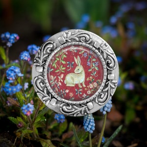 May include: A silver-toned metal pin with a round, glass-like center featuring a white rabbit on a red background with floral patterns. The pin has a detailed, ornate design around the edge.