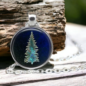 May include: A silver chain necklace with a round pendant featuring a blue background and a green and yellow silhouette of a pine tree.