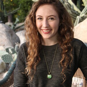May include: A woman with long, curly brown hair is wearing a dark gray sweater and a gold necklace with a green apple pendant. She is smiling and sitting in a wicker chair.