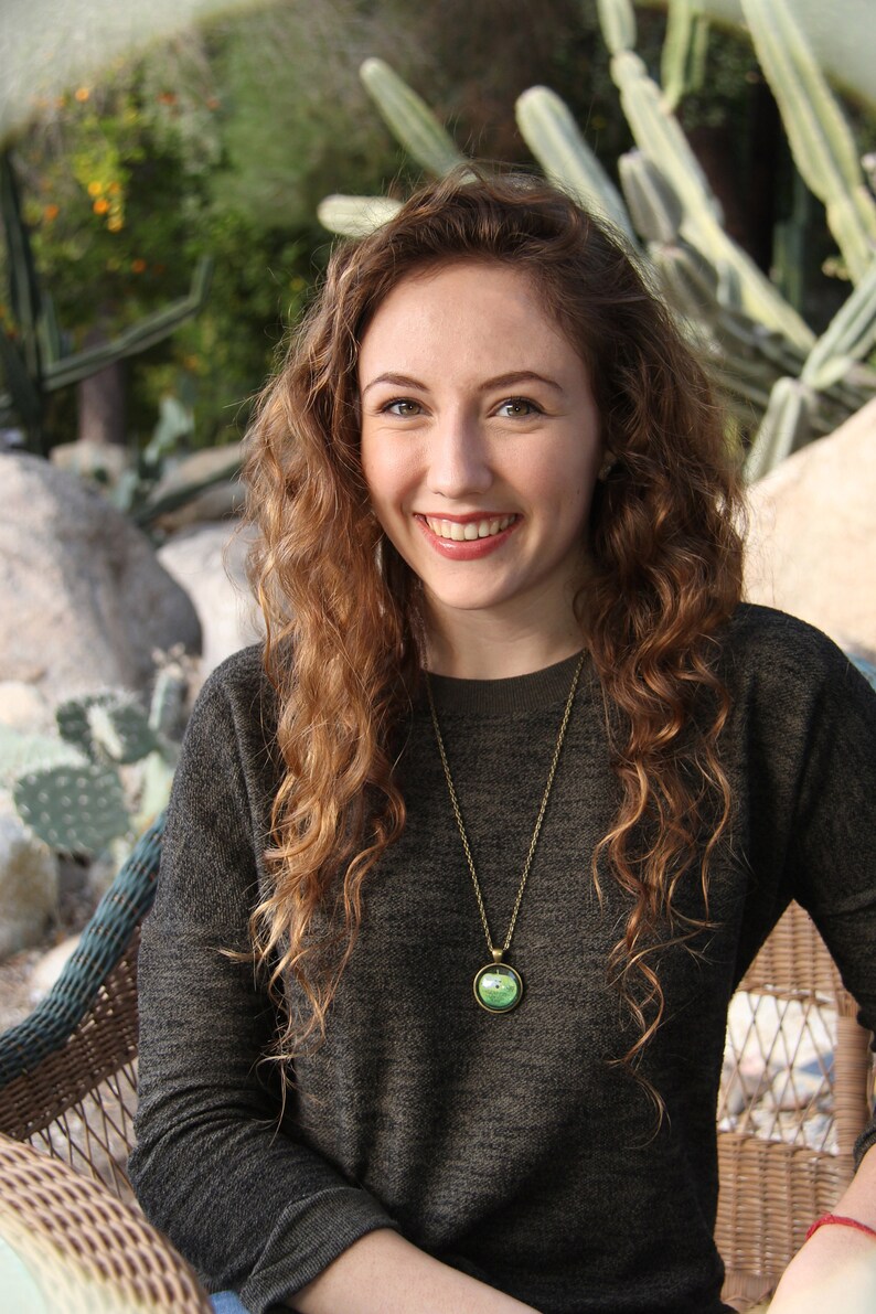 May include: A woman with long, curly brown hair is wearing a dark gray sweater and a gold necklace with a green apple pendant. She is smiling and sitting in a wicker chair.