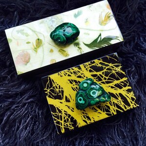 May include: Two small decorative boxes with a black background and a green malachite stone on top. One box has a white background with a floral pattern and the other has a yellow background with a black tree branch pattern.