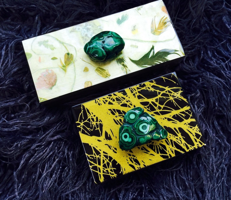 May include: Two small decorative boxes with a black background and a green malachite stone on top. One box has a white background with a floral pattern and the other has a yellow background with a black tree branch pattern.