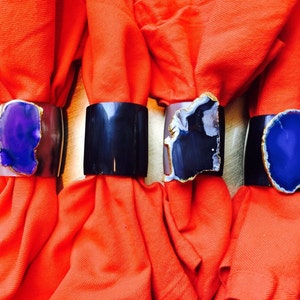 May include: Four napkin rings made of metal with a slice of agate stone in purple and black colours. The rings are on orange napkins.
