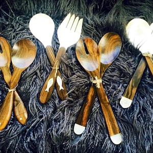 May include: Set of four wooden salad servers with a silver finish. Two servers have a spoon shape and two have a fork shape. The servers are on a black fuzzy surface.