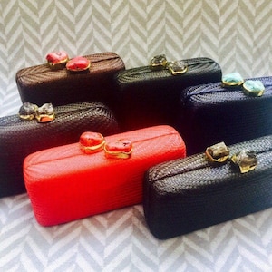May include: A collection of six woven clutches in various colours, each with a unique decorative clasp. The clutches are arranged in a row, with the brown clutch at the top and the black clutch at the bottom.
