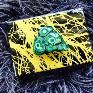 May include: A green malachite stone with a swirling pattern sits on a black and gold box with a tree branch design.