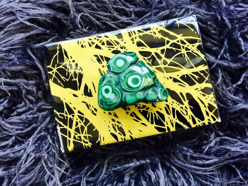 May include: A green malachite stone with a swirling pattern sits on a black and gold box with a tree branch design.