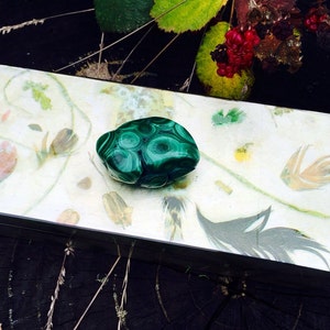 May include: A green malachite stone with swirling patterns sits on a decorative box with a white background and floral designs.