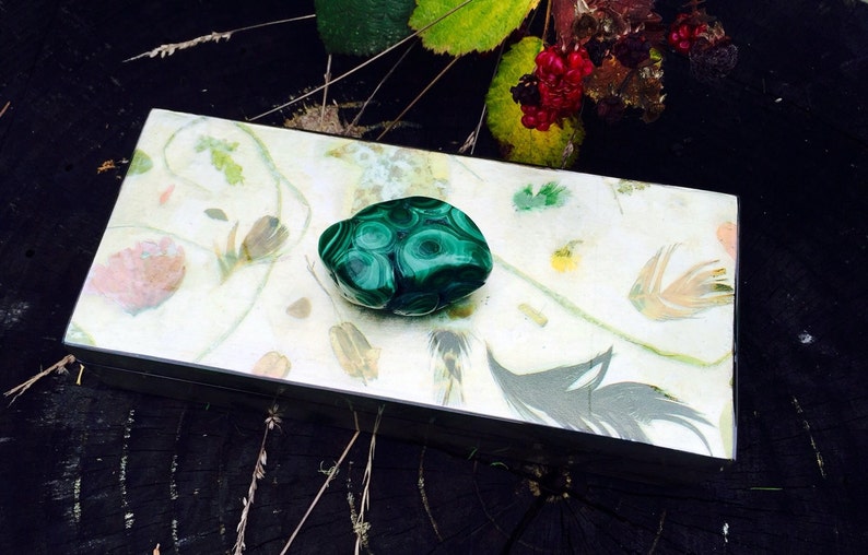 May include: A green malachite stone with swirling patterns sits on a decorative box with a white background and floral designs.