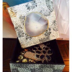 May include: A decorative box with a black and white patterned exterior. The lid is open and features a large, round, light gray agate slice with a gold rim. The interior of the box is black and contains a variety of decorative items.
