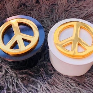 May include: Two round decorative boxes with a gold peace sign on the lid. One box is black and the other is white.