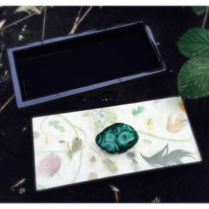 May include: A small black box with a green malachite stone inside. The box is open and the stone is sitting on a white background with a floral pattern.