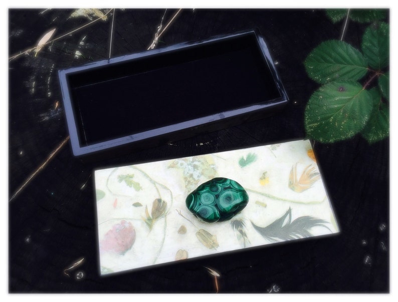 May include: A small black box with a green malachite stone inside. The box is open and the stone is sitting on a white background with a floral pattern.