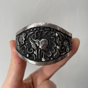 Könnte beinhalten: Ein silbernes Armband mit einem komplizierten floralen Design. Das Design ist erhaben und scheint handgeschnitzt zu sein.