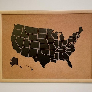 May include: A framed corkboard featuring a black outline of the United States map. The map is centered on the corkboard, with each state clearly defined. The frame is made of light-colored wood, and the background is a neutral color.