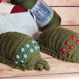 May include: Two handmade green crochet hats, one with blue beads and the other with red beads, sit on a wooden surface. A polka-dot fabric basket holds red yarn. Green yarn is also visible.