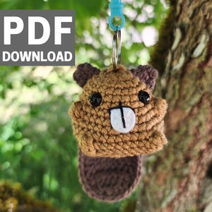 Op de afbeelding: Een handgemaakte gehaakte bever sleutelhanger in bruin- en paars tinten, met een witte tand en zwarte ogen. De sleutelhanger is bevestigd aan een blauwe plastic clip. De afbeelding bevat de tekst "PDF DOWNLOAD".