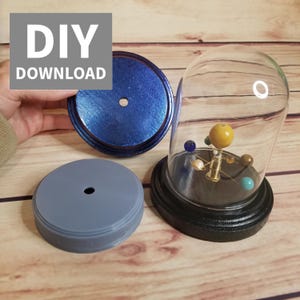 Op de afbeelding: DIY-zonnestelselmodelset met een glazen koepel, een houten basis en een blauwe en grijze basis. Het model heeft een gouden zon en planeten in verschillende kleuren.