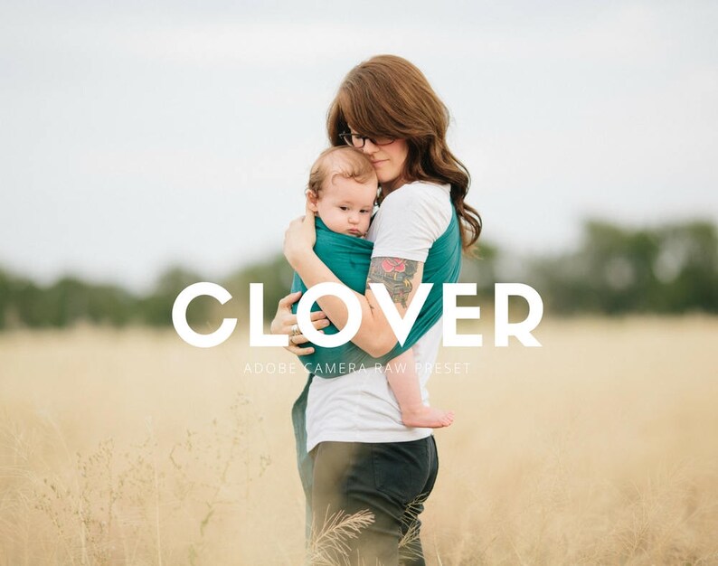 May include: A woman wearing a white shirt and a teal wrap carrier holds a baby in her arms. The woman has brown hair and is wearing glasses. The baby is wearing a teal wrap carrier and has brown hair. The background is a field of tall grass.