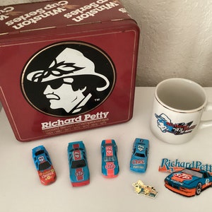 May include: A red metal box with a picture of Richard Petty and the text "Winston Cup Series" on the top. There are five toy race cars, a white coffee mug with a picture of Richard Petty, and a patch with the text "Richard Petty" on a white surface.