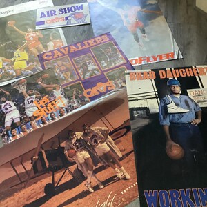 May include: A collection of vintage Cleveland Cavaliers basketball posters featuring players like Ron Harper, Brad Daugherty, and the team's logo. The posters are in various sizes and feature different designs, including a plane flying over a desert landscape and a basketball player in action.