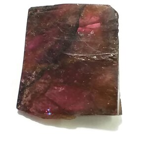 May include: A rough, dark pink and brown gemstone with a rectangular shape. The gemstone has a natural, unpolished finish.