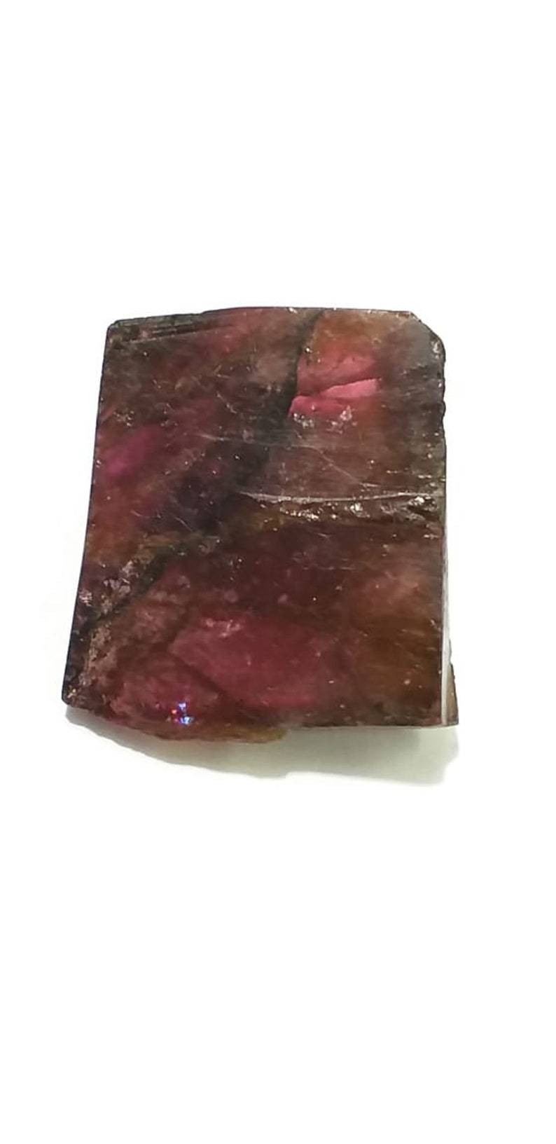May include: A rough, dark pink and brown gemstone with a rectangular shape. The gemstone has a natural, unpolished finish.
