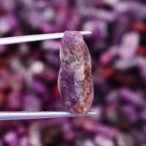 May include: A close-up of a purple rough ruby gemstone held by tweezers. The ruby has a striated texture and a deep purple color.