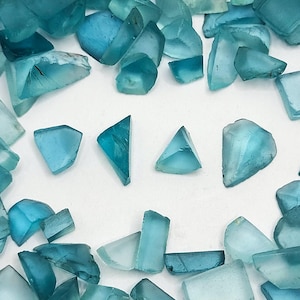 May include: A collection of teal blue gemstone chips, some are triangular, some are rectangular, and some are irregular shapes. The chips are scattered on a white surface.
