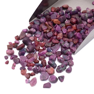 Può includere: Un mucchio di piccole pietre preziose ruvide, di colore viola e rosso. Le pietre preziose sono sparse su una superficie bianca e si riversano da una paletta di metallo.