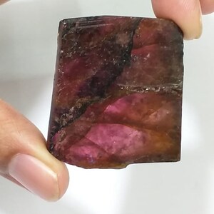 May include: A rough, rectangular, pink and brown gemstone with a dark brown vein running through it.