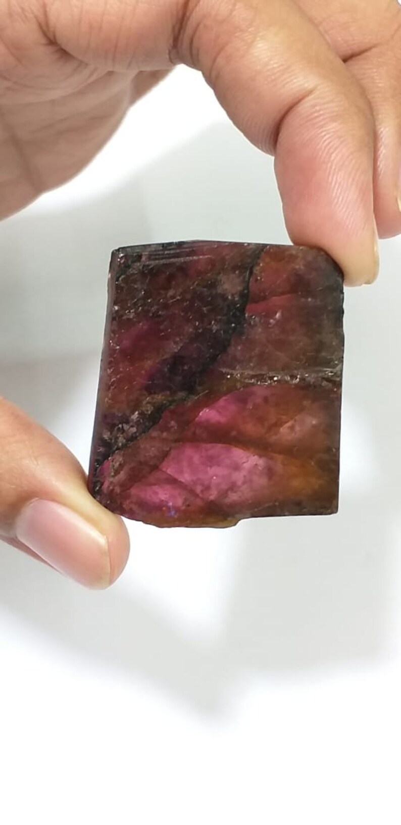 May include: A rough, rectangular, pink and brown gemstone with a dark brown vein running through it.