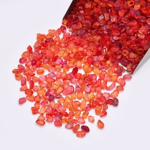 May include: A pile of loose red and orange gemstones, some with a more yellow hue, scattered on a white surface. The gemstones are various sizes and shapes, with some appearing more rounded and others more angular.