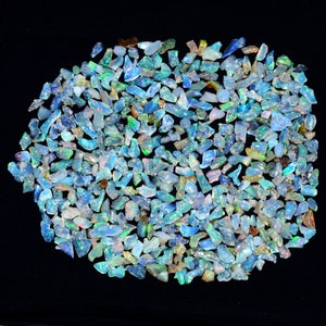 May include: A large collection of small, iridescent opal chips in shades of blue, green, and white. The chips are scattered on a black background.