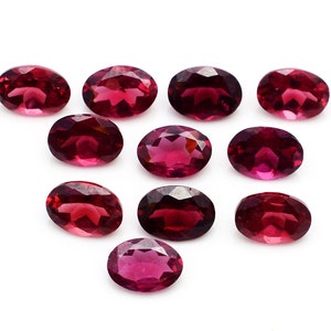 Puede incluir: Diez piedras preciosas de forma ovalada y facetadas en varios tonos de rojo. Las piedras preciosas están dispuestas en forma triangular sobre un fondo blanco.
