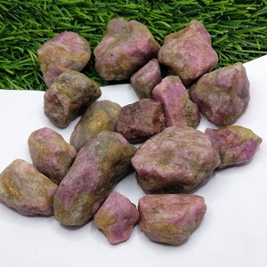 May include: A collection of rough, uncut ruby gemstones. The stones display a mix of pink, red, and brown hues, with a natural, unpolished appearance. The gemstones are scattered on a white surface with a green background.